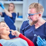 A hygienist smiles at his patient while a nurses preps in the background.