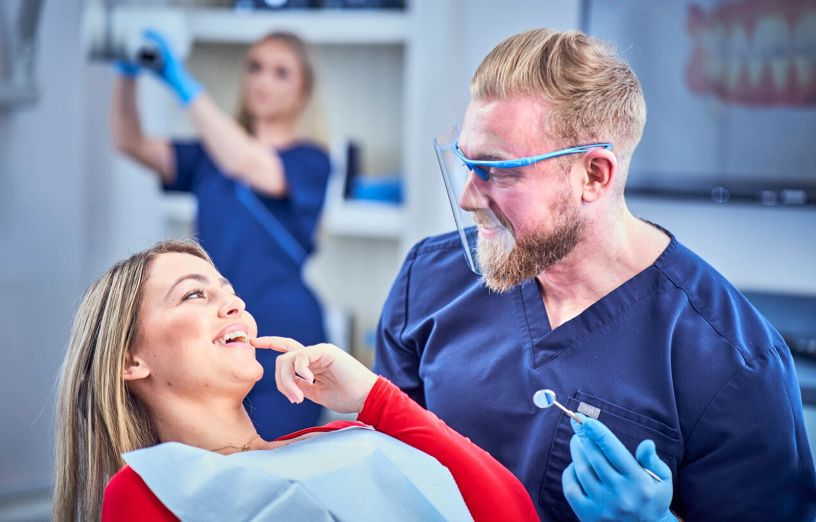 A hygienist smiles at his patient while a nurses preps in the background.
