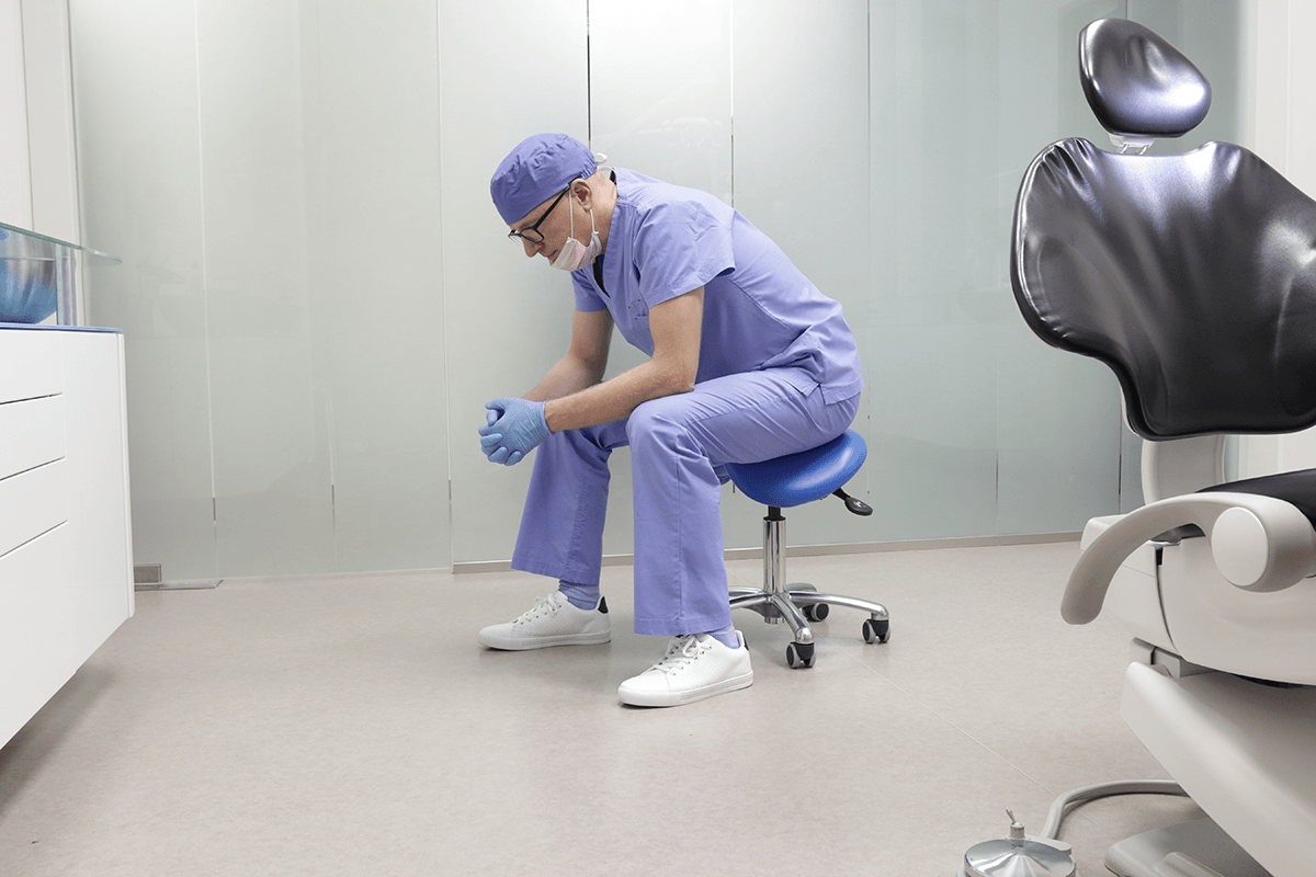 A dentist sits on a stool in his surgery, hunched over looking worried