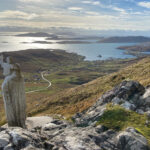 Serene coastal landscape with a light stone statue raising a hand on a rocky outcrop, overlooking sunlit water, distant islands, and a winding road through scattered hillside homes under partly cloudy skies.