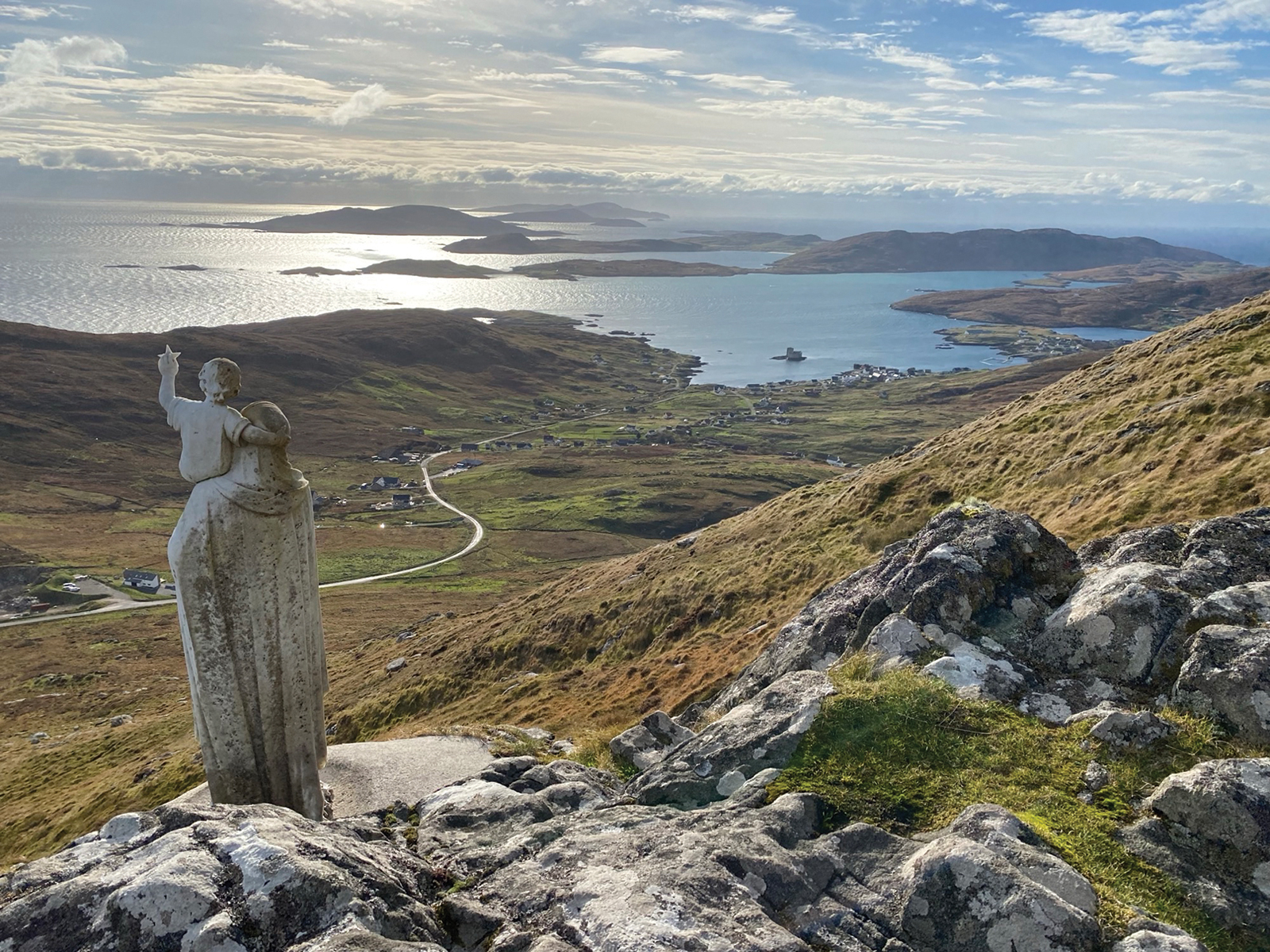 Serene coastal landscape with a light stone statue raising a hand on a rocky outcrop, overlooking sunlit water, distant islands, and a winding road through scattered hillside homes under partly cloudy skies.