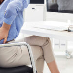 A woman, at her desk, rubs her lower back indicating pain.