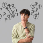 A man poses like he's thinking, a grey wall behind is filled with black line drawn question marks.