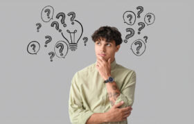 A man poses like he's thinking, a grey wall behind is filled with black line drawn question marks.