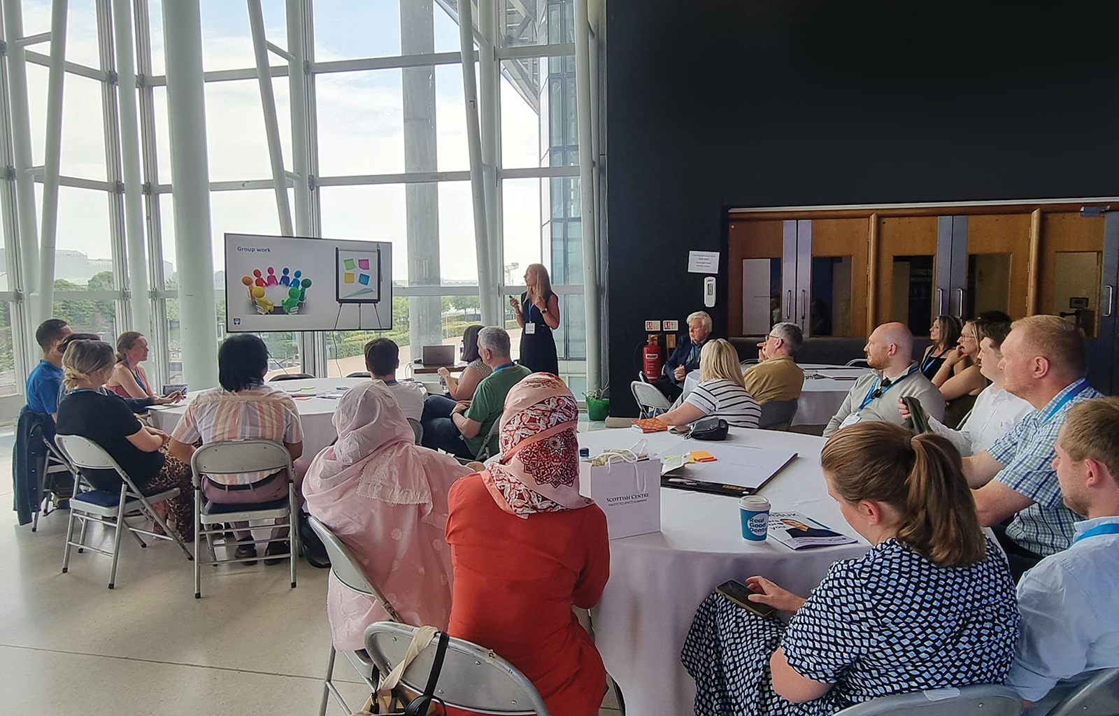 A blonde woman speaks too an audience sitting at three large round tables. They are in a bright glass walled room, looking towards a large screen.