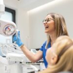 A dentist adjusts the overhead spotlight while laughing with her patient.