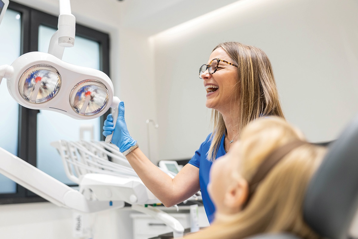 A dentist adjusts the overhead spotlight while laughing with her patient.