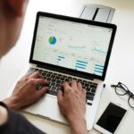 A man looks at some charts and graphs on his laptop, his mobile phone and glasses sit to the right of the laptop.