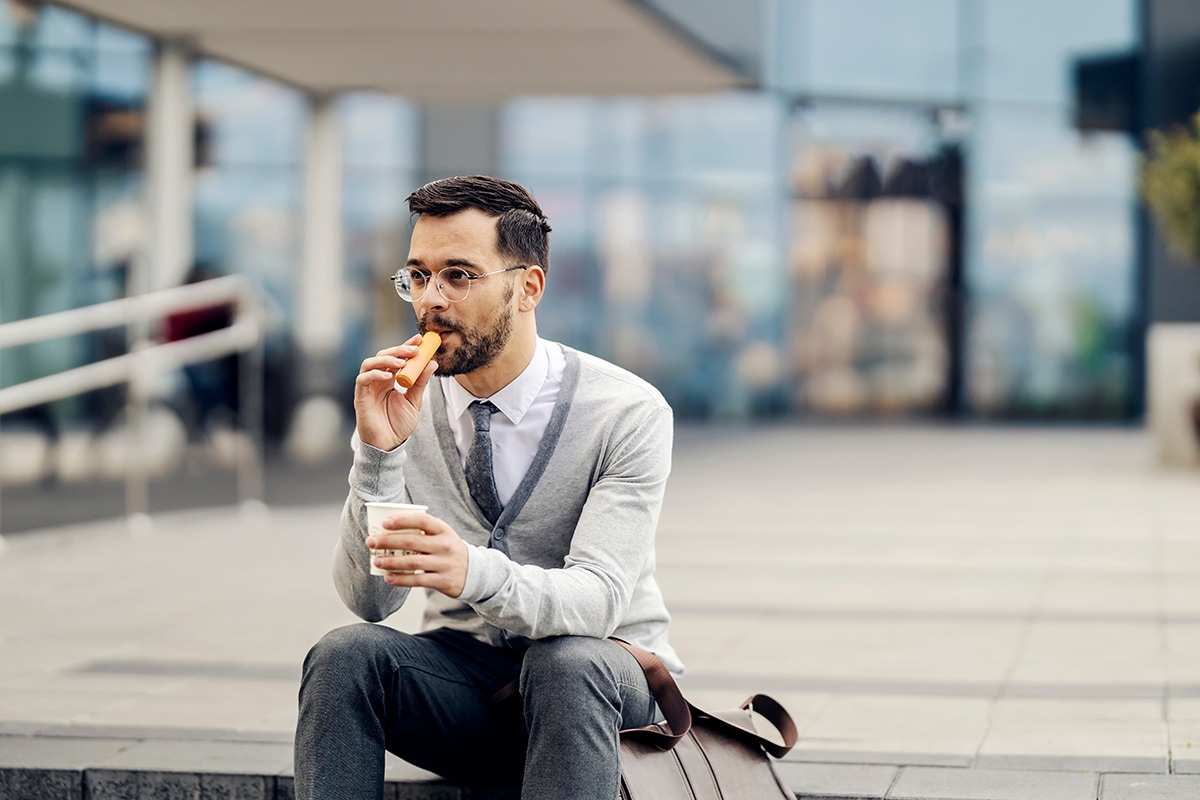 A well dressed man sits on a step outside a glass office block, he's holding a coffee cup and smoking from an electronic vape pen.