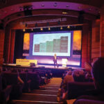 Professor Kebschull walks across stage in a darkened theatre.