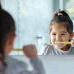 A little girl stands and brushes her teeth looking in a mirror.