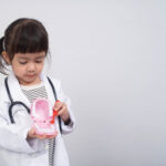 A little girl in a lab coat wearing a toy stethoscope brushes a set of toy teeth