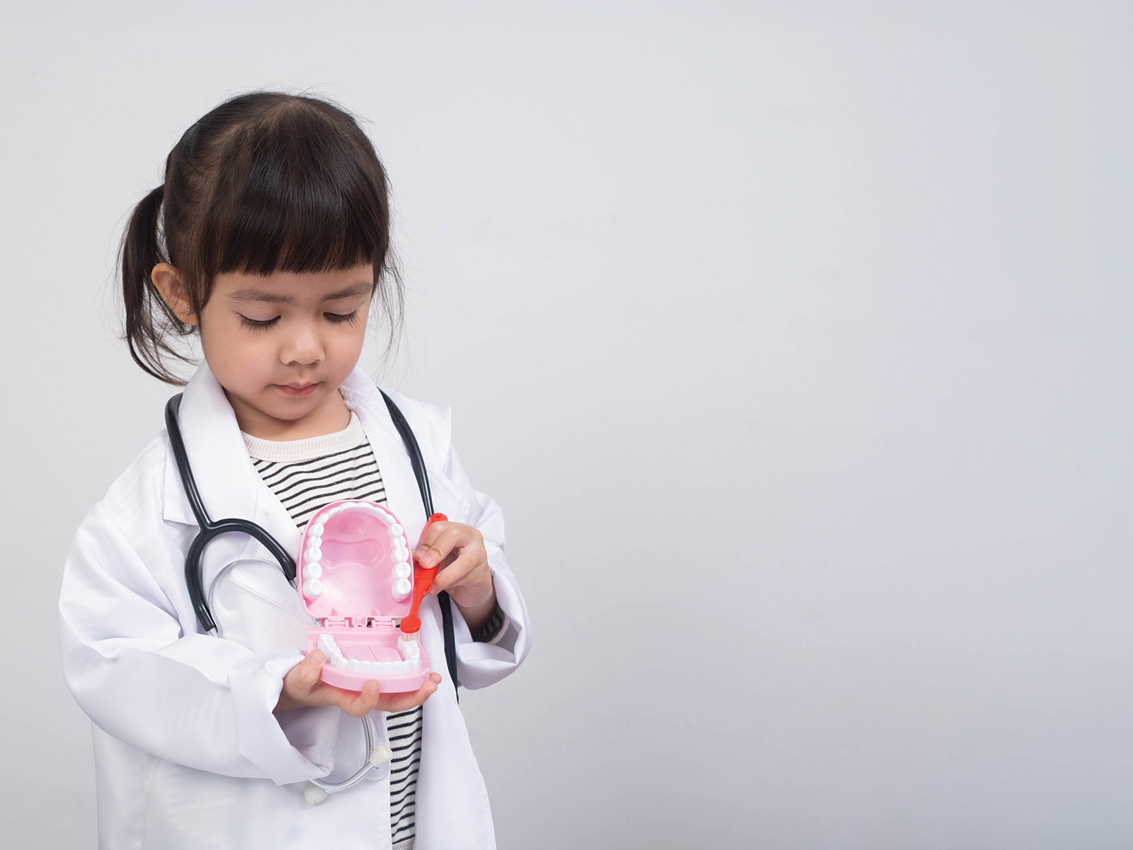 A little girl in a lab coat wearing a toy stethoscope brushes a set of toy teeth